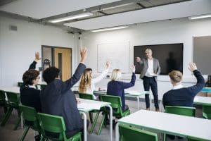 Rear view of engaged teenage students in uniform sitting at desks, mature male teacher standing in front and pointing to boy raising his hand.